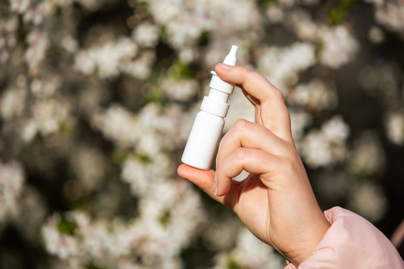 woman's hand holding allergy nasal spray in front of white flowers
