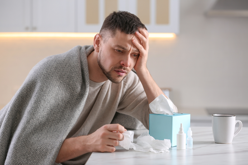 man with nasal congestion sitting at table in front of tissues, nasal spray, and cup of hot tea